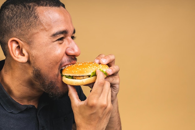 Hungry young african american black man eating hamburger isolated over beige background Diet or Fast food concept