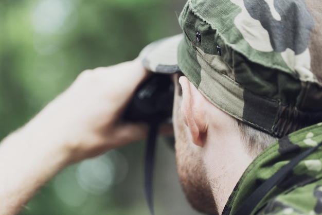 hunting, war, army and people concept - close up of young soldier, ranger or hunter with binocular observing forest