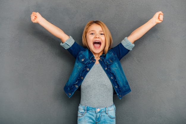 Photo hurray! cheerful little girl keeping arms outstretched and looking at camera while standing against grey background