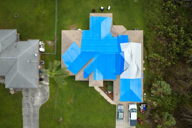 Hurricane Ian damaged house rooftop covered with protective plastic tarp against rain water leaking until replacement of asphalt shingles Aftermath of natural disaster