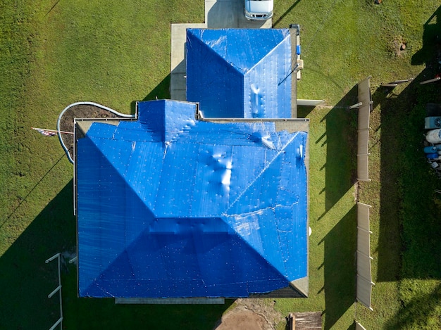 Hurricane Ian damaged house rooftop covered with protective plastic tarp against rain water leaking until replacement of asphalt shingles Aftermath of natural disaster