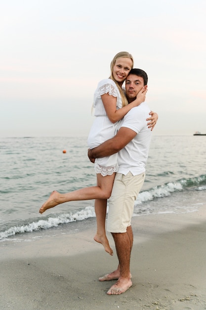 husband holding his wife on the beach near the ocean at sunset. romantic couple