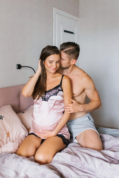 Husband hugs his pregnant wife while sitting on the bed in the morning