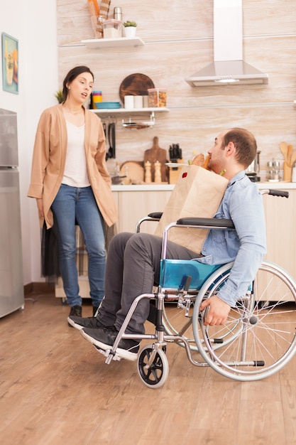 Husband in wheelchair with paper bag from supermarket in kitchen talking with wife. Disabled paralyzed handicapped man with walking disability integrating after an accident.