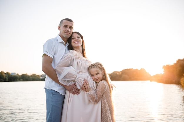 A husband with a pregnant wife and daughter is resting on the river bank standing on the pier in summer sunny weather at sunset couple in love man and woman hugging holding hands A happy family