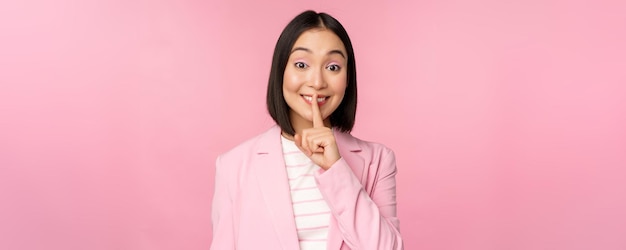 Hush taboo concept Portrait of asian businesswoman showing shush gesture shhh sign press finger to lips standing over pink background in suit