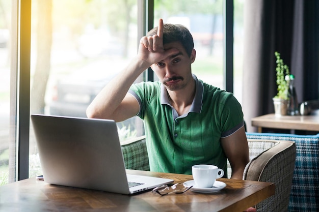 I am loser Young sad businessman in green tshirt sitting and looking at camera with loser gesture on his forehead and feel sad business and freelancing concept indoor shot near window at daytime