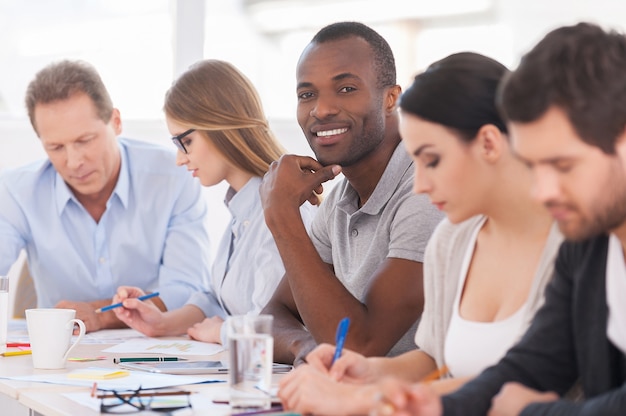 I am a part of strong and creative team. Group of business people sitting in a row at the table while handsome African man looking at camera and smiling