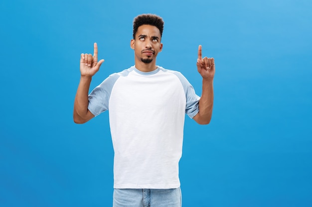 I am unsure about it. Portrait of uncertain and doubtful dark-skinned man with beard and afro hairstyle frowning and smirking suspiciously looking and pointing up hesitating from hard choice