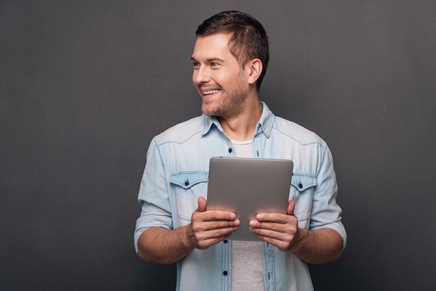 I can solve your problem! Cheerful young man holding his digital tablet and looking away with smile while standing against grey background