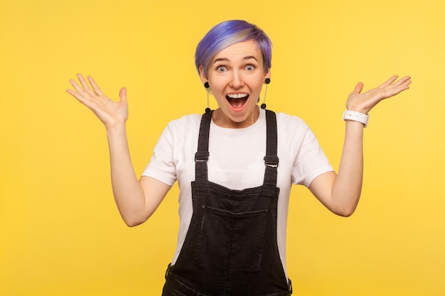 I can't believe this. Portrait of amazed hipster girl with violet short hair in denim overalls raising hands from sudden surprise, awesome shocking news. isolated on yellow background, studio shot