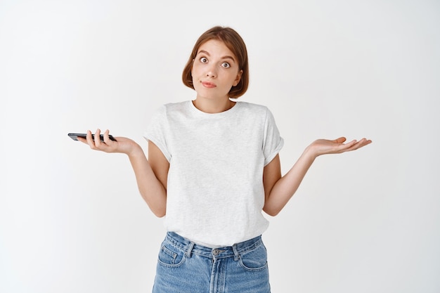 I dont know. Clueless european woman shrugging shoulders, have no idea, holding mobile phone and look questioned, standing against white wall