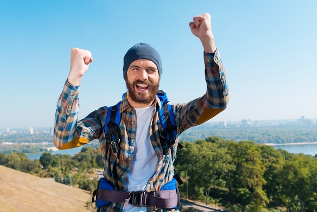 I have made it! Happy young man carrying backpack and looking at camera with raising arms