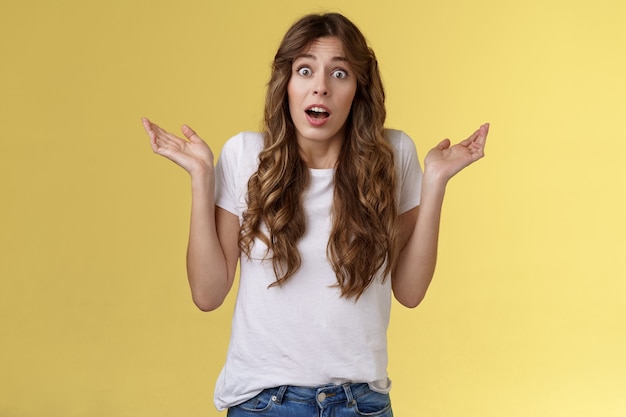 I not know. Clueless confused cute nervous young woman shrugging hands raised sideways full disbelief questioned expression lift eyebrows puzzled unable answer question stand yellow background