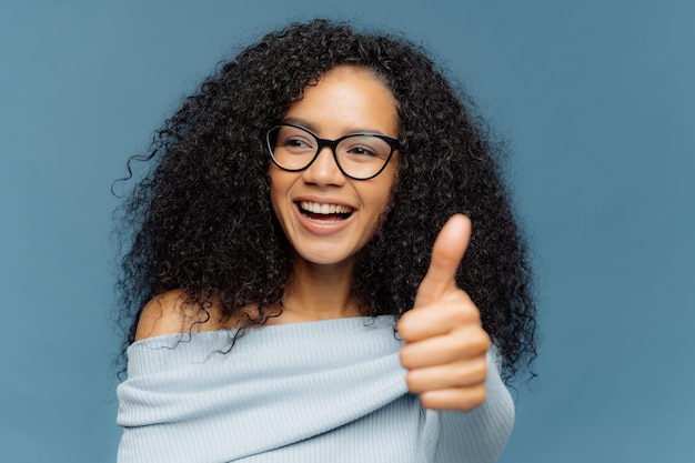 I like it Portrait of happy woman with Afro hairstyle shows thumb up gives positive opinion looks aside wears optical glasses and sweater isolated on blue background People agreement approval