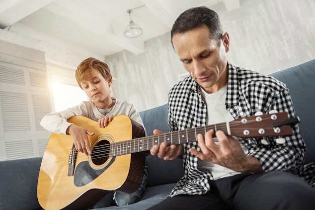 I like playing. Attractive concentrated little fair-haired holding the guitar and his father teaching him to play the guitar and they sitting on the couch