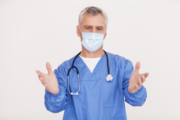 I will help you! Portrait of senior grey hair doctor in surgical mask looking at camera and gesturing while standing isolated on white