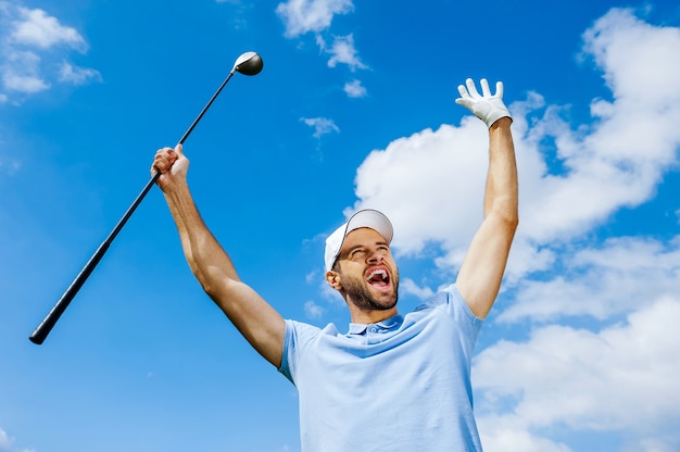 Photo i won! low angle view of young happy golfer holding driver and raising his arms with blue sky as background