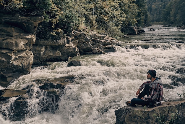 I would stay here forever. Handsome young modern man looking away while sitting on the rock near the river