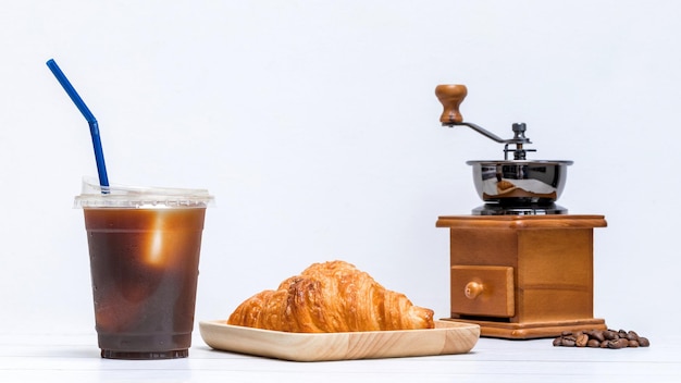 Ice americano coffee in a cup with croissant on wooden plate and coffee bean grinder ready for breakfast meal