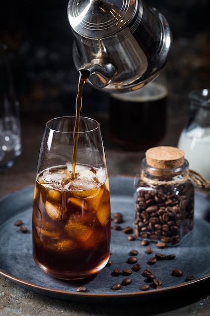 Ice coffee cold summer drink in a tall glass with pouring coffee and coffee beans on a stone background.