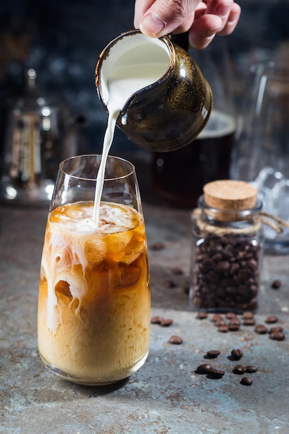 Ice coffee cold summer drink in a tall glass with pouring cream and coffee beans on a stone background.