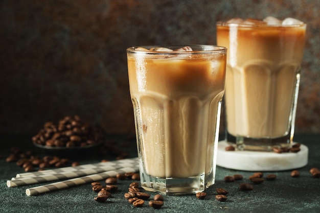 Ice coffee in a tall glass with cream poured over ice cubes and beans on a dark concrete table Cold summer drink with tubes on a black background with copy space