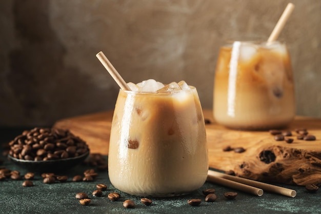 Ice coffee in a tall glass with cream poured over ice cubes and beans on a dark concrete table Cold summer drink with tubes on a black background with copy space