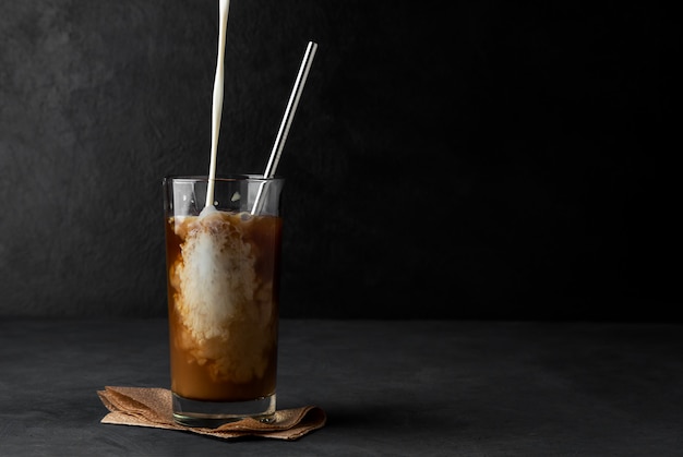 Iced coffee with pouring cream in a glass cup with a metal straw on a dark space