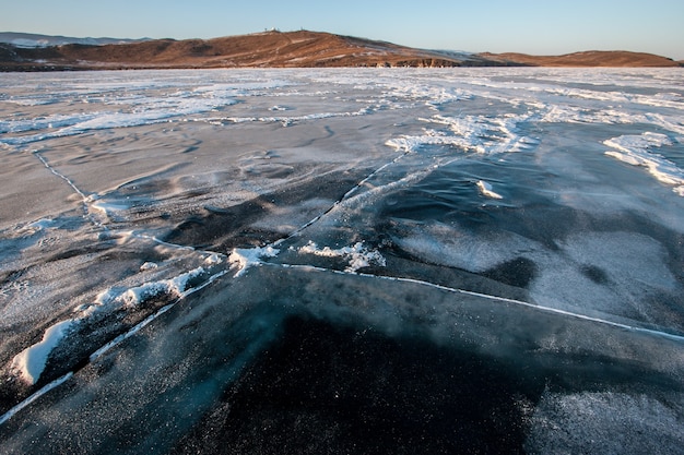 The icy surface of Lake Baikal in winter with large cracks, snow and hills in the background. The thickness of the ice is visible. Sunny weather, clear sky.