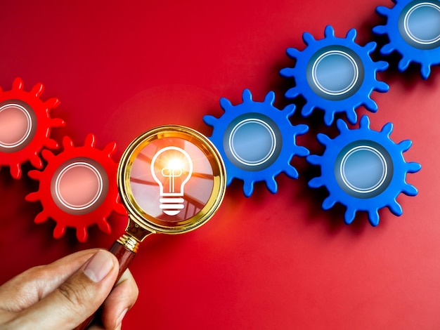 Idea light bulb symbol appear on magnifying glass lens in hand appear between blue and red gears on red background Business strategy creative thinking solution human research teamwork concepts