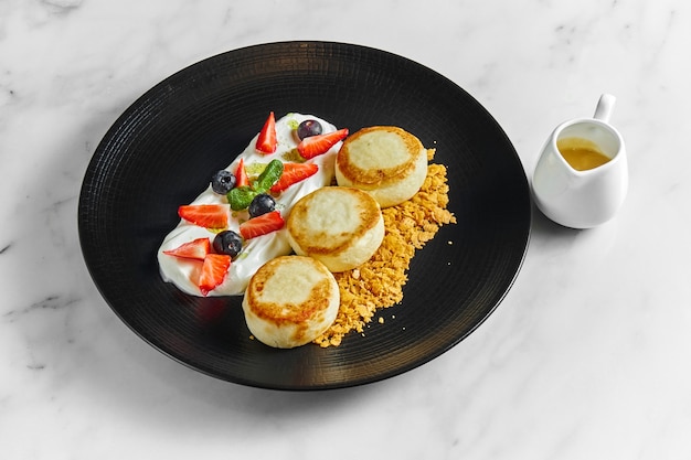 An ideal breakfast - Cottage cheese pancakes with white sauce, berries, strawberries and crunch in a black plate on a marble surface