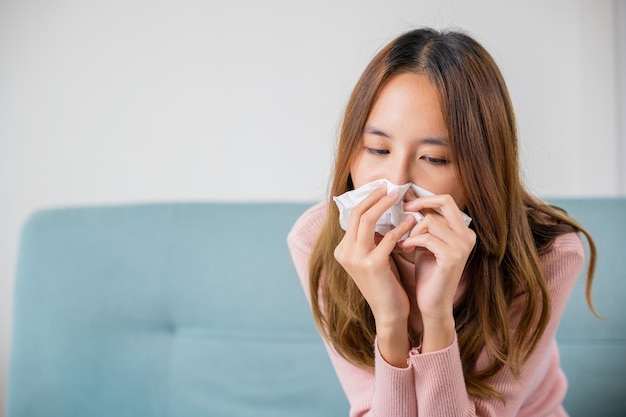 Ill Asian young woman cold covered with blanket sitting on sofa and sneeze with tissue paper at home, Female sick allergic blowing nose sneezing in tissue at home, Flu health care