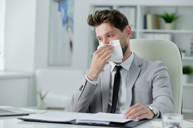 Ill young businessman sitting at desk and rubbing runny nose while working with papers in office, cold and flu concept