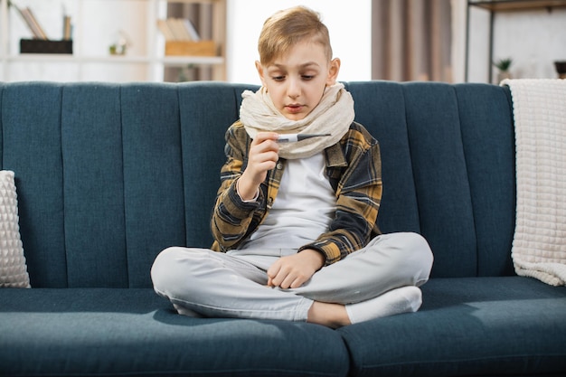 Illness flu and cold concept Little sick teen boy sitting in couch checking temperature