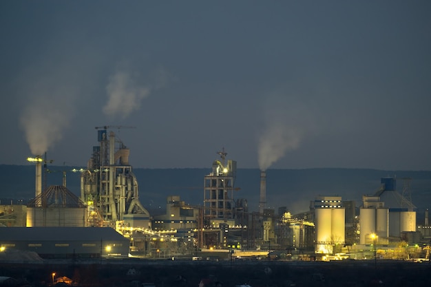 Illuminated cement plant with high factory structure and tower cranes at industrial production area at night Manufacture and global industry concept