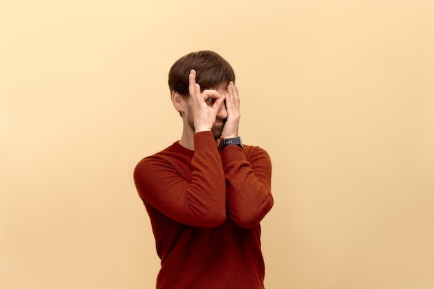 Im here, im fine. Photo of funny young man with beard wearing sweater, make okay gesture over eye, covered another half face with palm, posing against beige wall.