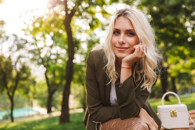 Image of adorable woman 20s smiling, while sitting on bench in park with trees