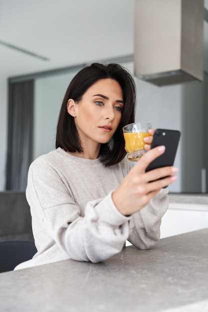 Image of adult woman 30s drinking orange juice and using mobile phone, while resting in bright modern room