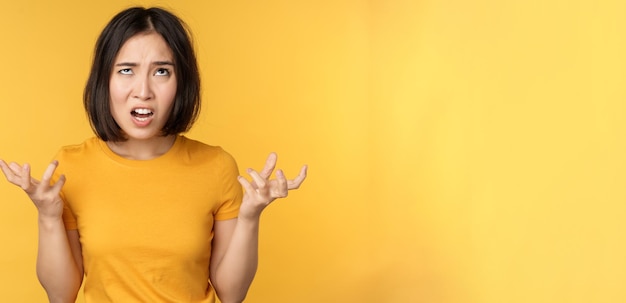 Image of angry asian woman shouting and cursing looking outraged furious face expression standing over yellow background