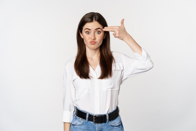 Image of annoyed young woman showing finger gun gesture over head, bothered by smth, tired, standing over white background.