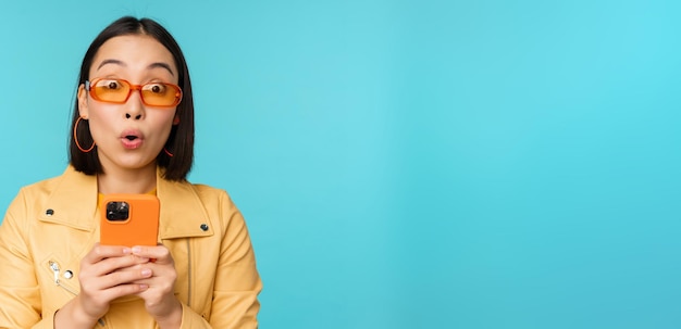 Image of asian girl in sunglasses looking amazed and impressed recording video or taking photo on smartphone standing over blue background