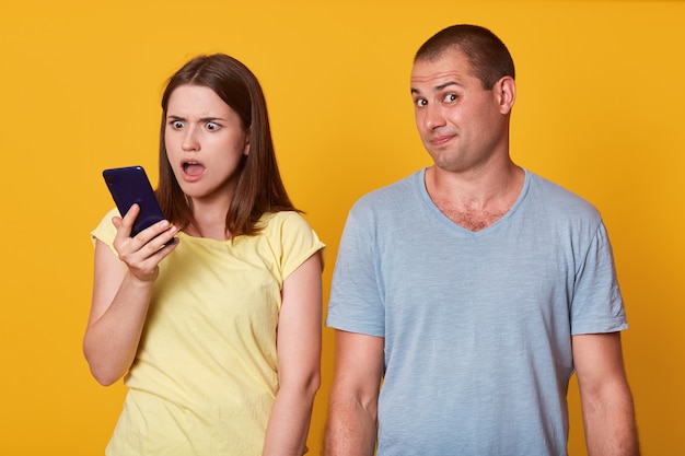 Image of astonished woman holding husband's phone, checking his messages with open mouth and shocked facial expression