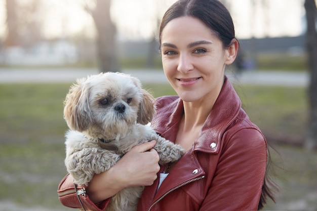 Image of attractive charismatic brunette looking directly at camera, having pleasant facial expression, holding her Maltese dog, being fond of animals, enjoying spring time.