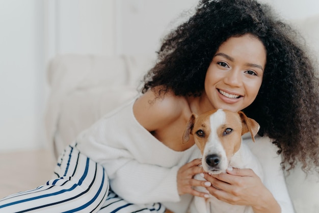 Image of attractive dark skinned woman wears makeup has bushy curly hair smiles pleasantly cuddles dog dressed in fashionable clothes enjoys sweet moment with puppy being at home together