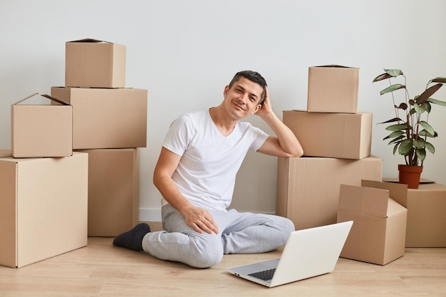 Image of attractive smiling man wearing white t shirt sitting on floor surrounded with cardboard boxes with belongings and posing in front of laptop expressing positive emotions