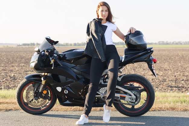 Image of attractive woman wears white t shirt, black trousers and white trainers, sits on motorrbike, keeps hand on helmet