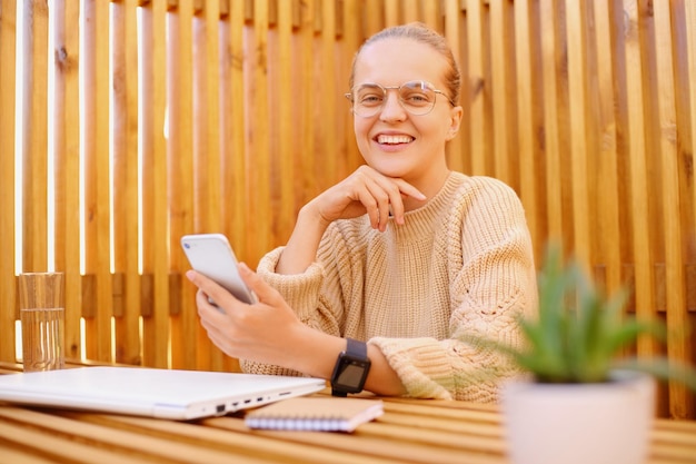 Image of attractive young adult woman with bun hairstyle wearing beige sweater sitting at table with closed laptop and holding smart phone looking smiling at camera