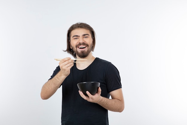 Image of bearded man holding bowl with chopsticks over a white wall.