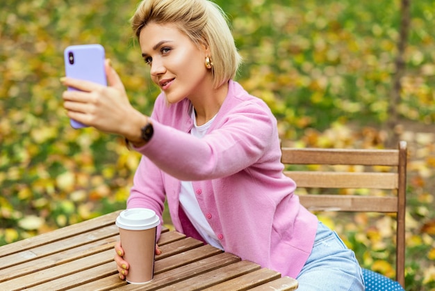 Image of beautiful blonde young woman wearing blue jeans white tshirt and pink jacket smiling and taking selfie on smartphone Happy female making self portrait in the city park